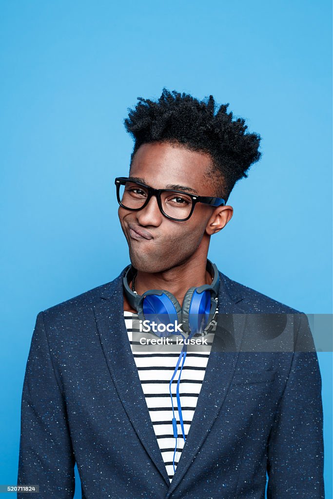 Funky afro american guy against blue background Studio portrait of fashionable afro american young man wearing striped top, navy blue jacket, nerd glasses and headphone, making face. Studio portrait, blue background. Asking Stock Photo