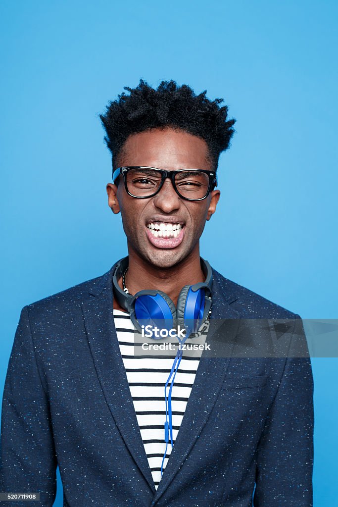 Funky afro american guy clenching teeth Studio portrait of fashionable afro american young man wearing striped top, navy blue jacket, nerd glasses and headphone, clenching teeth. Studio portrait, blue background. Eyeglasses Stock Photo