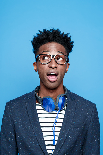 Studio portrait of surprised afro american young man wearing striped top, navy blue jacket, nerd glasses and headphone, looking up with mouth open. Studio portrait, blue background.