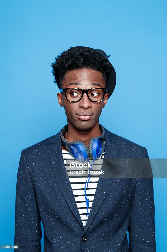 Fashionable afro american young man against blue background Studio portrait of pensive afro american young man wearing striped top, navy blue jacket, nerd glasses, hat and headphone, looking away. Studio portrait, blue background. Portrait Stock Photo