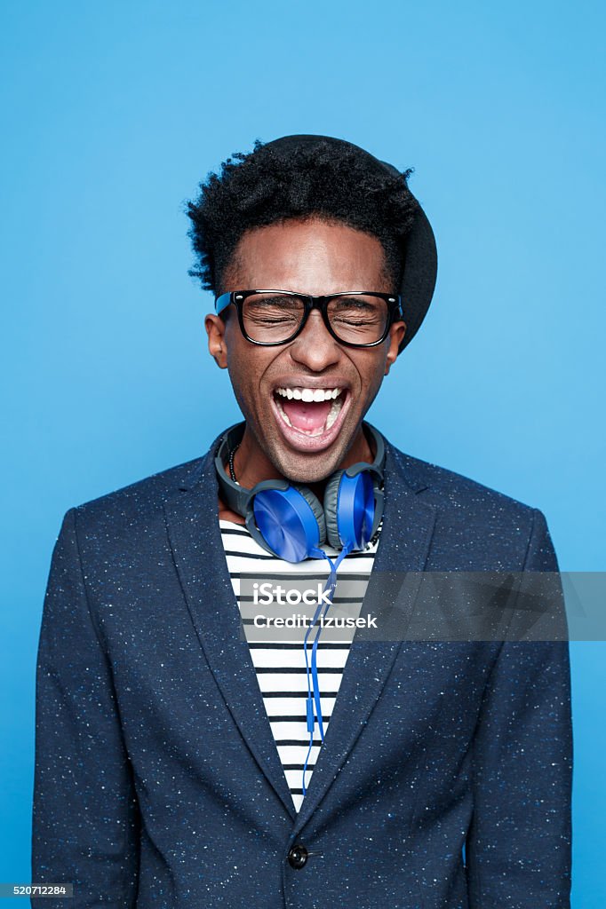 Funky afro american guy screaming Studio portrait of angry afro american young man wearing striped top, navy blue jacket, nerd glasses, hat and headphone, shouting. Studio portrait, blue background. Colored Background Stock Photo