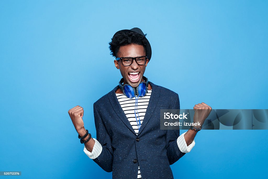 Fashionable afro american guy expressing happiness Studio portrait of successful afro american young man wearing striped top, navy blue jacket, nerd glasses, hat and headphone, laughing at camera with raised fists. Studio portrait, blue background. Men Stock Photo