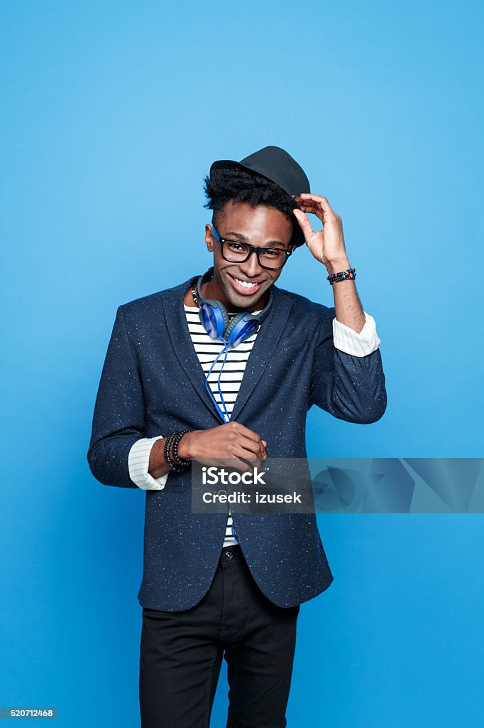 Funky afro american guy in fashionable outfit Studio portrait of successful afro american young man wearing striped top, navy blue jacket, nerd glasses, hat and headphone, smiling at camera. Studio portrait, blue background. Greeting Stock Photo