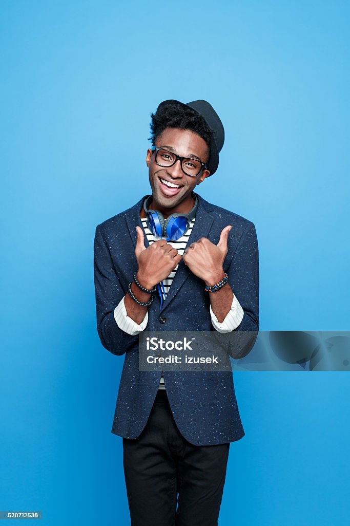 Funky afro american guy in fashionable outfit Studio portrait of successful afro american young man wearing striped top, navy blue jacket, nerd glasses, hat and headphone, smiling at camera. Studio portrait, blue background. Men Stock Photo