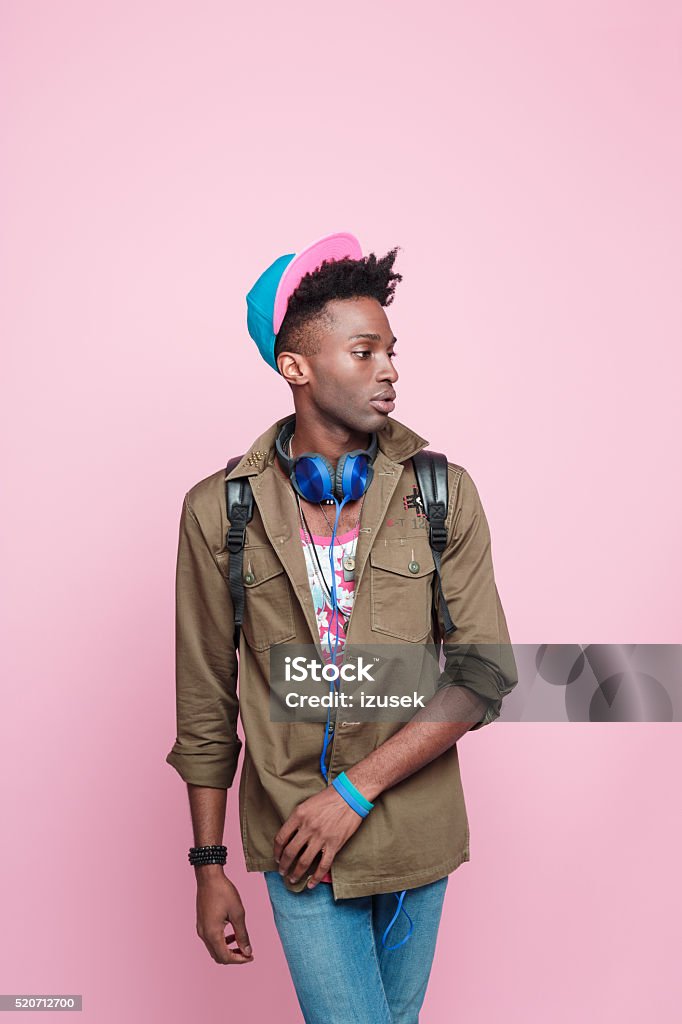 Studio portrait of carefree afro american student Summer portrait of cool, fashionable afro american young man in modern outfit, wearing headphone, cap, jacket and backpack, standing against pink background. Adult Stock Photo