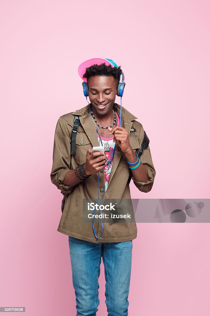 Happy afro american student using smart phone, studio shot Studio portrait of cute afro american young man wearing headphone, standing against pink background and using smart phone. Headphones Stock Photo