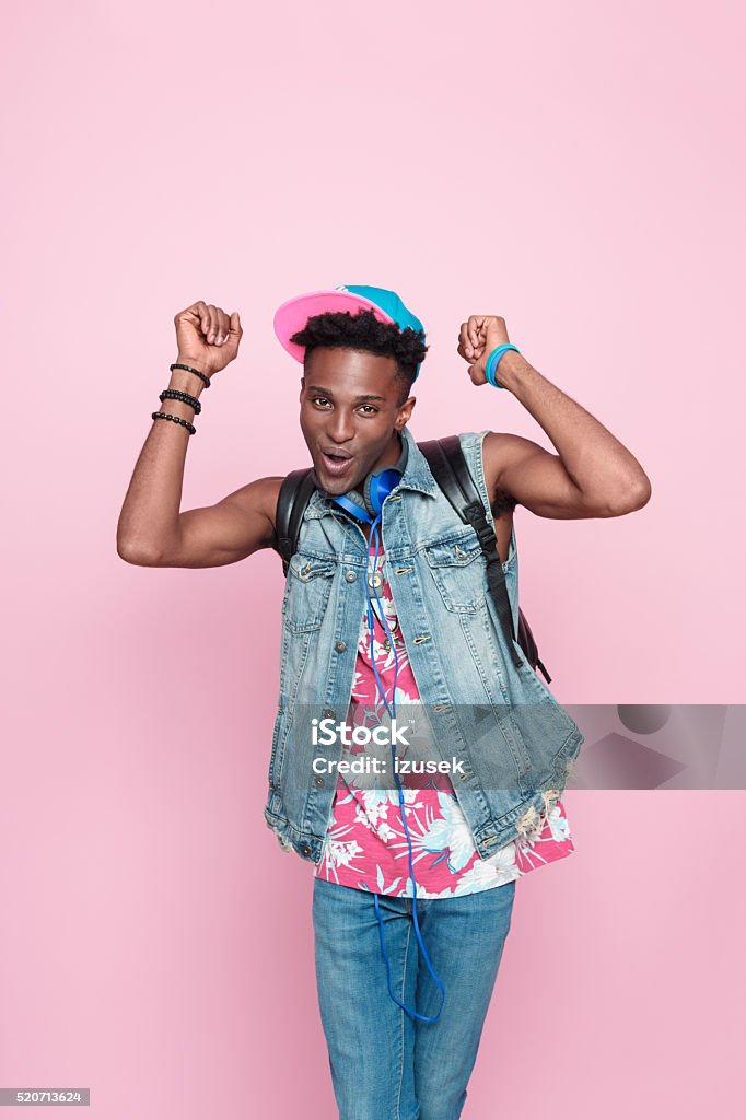 Summer portrait of funky afro american guy Summer portrait of happy afro american young man wearing headphone, cap and jeans sleeveless jacket, standing against pink background, laughing at camera with raised hands. Backpack Stock Photo