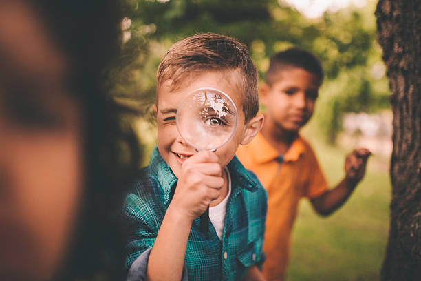 Boy in park holding a magnifying glass to his eye Portrait of a little boy holding up a large round magnifying glass to his face, making his eye look humourously large, while playing with friends in a summer park magnifying glass photos stock pictures, royalty-free photos & images
