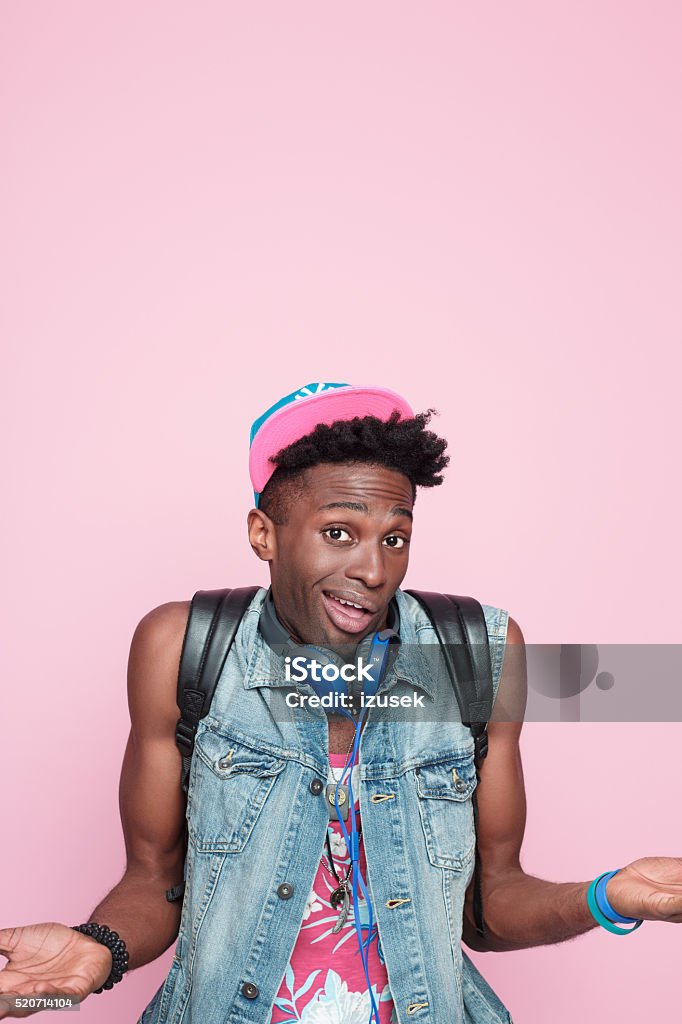 Summer portrait of surprised afro american guy Summer portrait of surprised afro american young man wearing headphone, cap and jeans sleeveless jacket, standing against pink background, looking at camera. Adult Stock Photo
