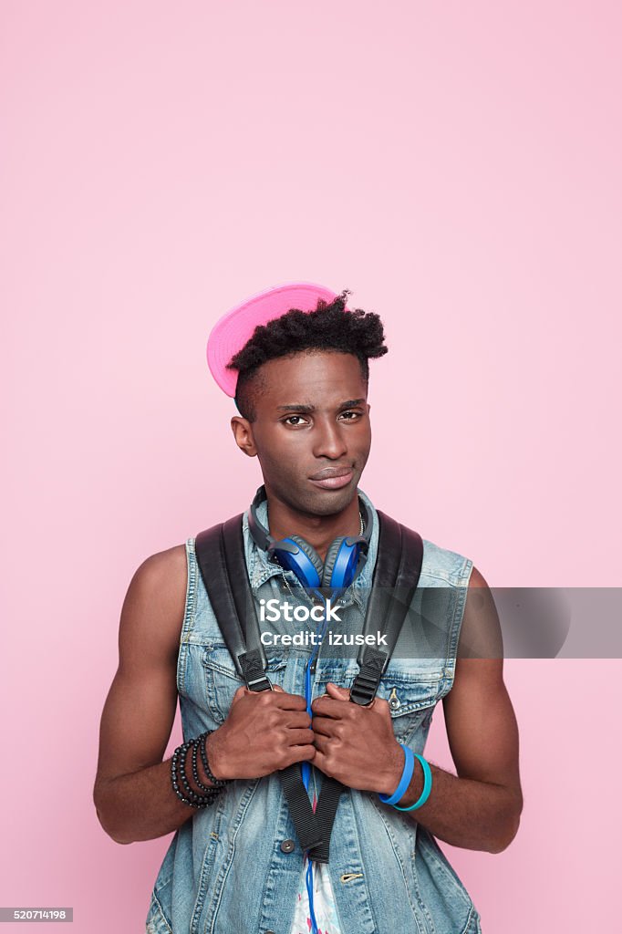 Summer portrait of disappointed afro american guy Summer portrait of disappointed afro american young man wearing headphone, cap and jeans sleeveless jacket, standing against pink background, looking at camera. Adult Stock Photo