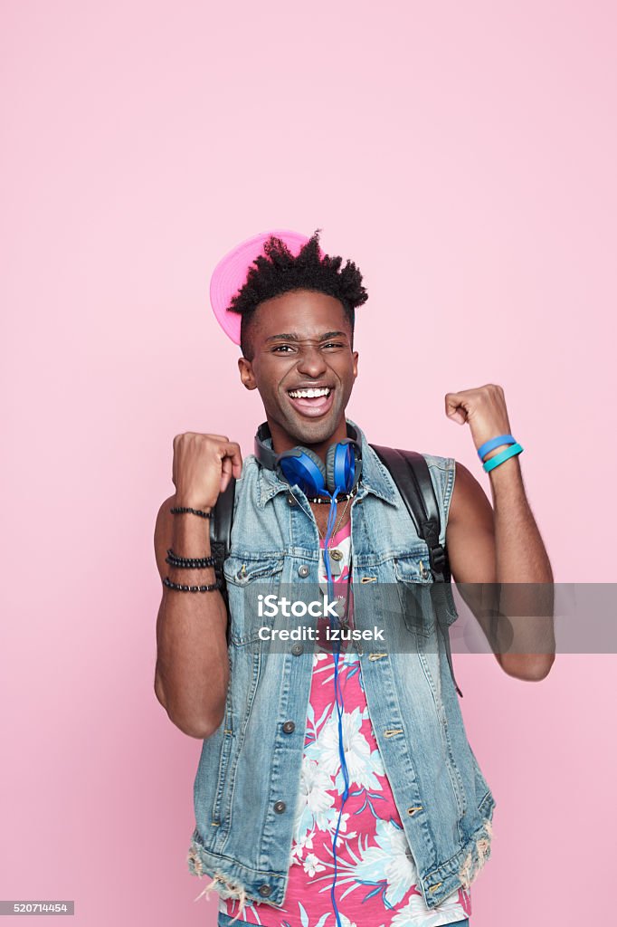 Summer portrait of excited afro american guy Summer portrait of happy afro american young man wearing headphone, cap and jeans sleeveless jacket, standing against pink background, laughing at camera with raised hands. Adult Stock Photo