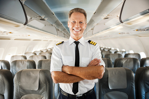 Confident male pilot in uniform keeping arms crossed and smiling while standing inside of the airplane