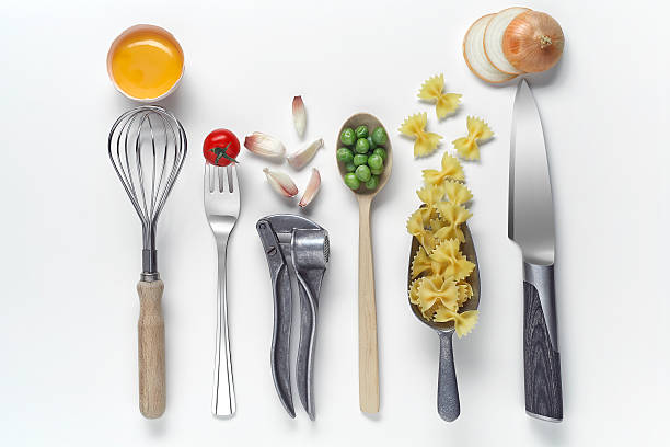 Top view shot of practical kitchen utensils and fresh cooking ingredients which are interrelated and aligned on white background. There is a harmony between wire whisk and egg, fork and tomato, garlic press and garlic piece, wooden spoon and green pea, metal measuring spoon and pasta, kitchen knife and onion slices.