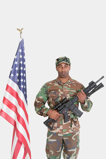 Portrait of US Marine Corps soldier with M4 assault rifle standing by American flag over gray background