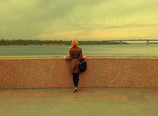 Beautiful young woman is looking at view over the river rear shot. Attractive girl enjoys summer and warm sunlight. The wind blew her hair. Vintage color, copyspace