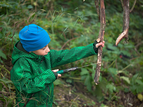 Little boy in a cap and jacket handsaw sawing dry snag on grass background
