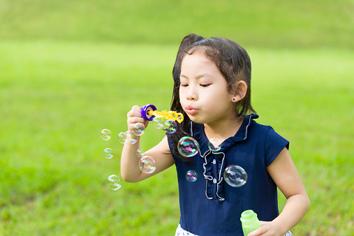 Playful 6 years old Asian Chinese Girl in navy blue and white dress, blowing bubbles with a bubble wand at a field.