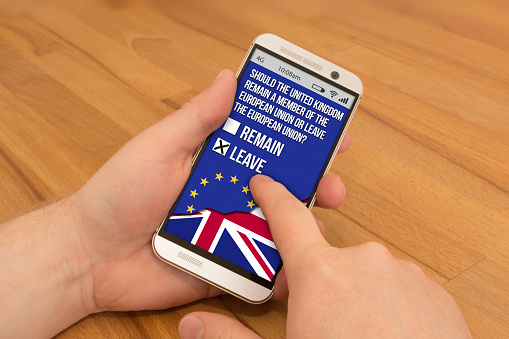Holding a smartphone in one hand, swiping or using gesture control with the other hand. On the screen is showing the EU Referendum question the UK will vote on. Leave is selected.