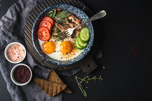 Breakfast set. Pan of fried eggs with bacon, fresh tomato, cucumber, sage and bread on dark serving board over black background, top view, copy space