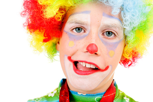 Portrait of a grimacing boy with his face painted as clown, wearing a multi colored wig. White background.