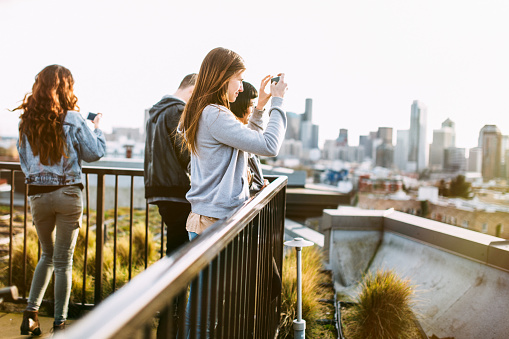 A happy group of young adults have fun together on a rooftop over looking downtown Seattle at dusk, taking pictures of the cityscape.  They wear trendy clothing, representative of Capitol Hill and city culture. Horizontal image with copy space.