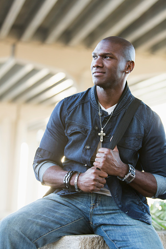 Portrait of a mixed race African American and Hispanic mid adult man with a serious expression, wearing jewelry and denim. He has an earring and a cross on a chain around his neck.