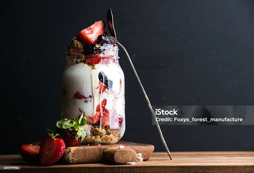 Yogurt oat granola with strawberries, mulberries, honey and mint leaves Yogurt oat granola with strawberries, mulberries, honey and mint leaves in tall glass jar on black backdrop, selective focus, copy space Dessert - Sweet Food Stock Photo