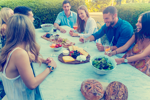 Group of young people eating outdoors. They are all happy, having fun, smiling laughing and talking. There are fruit and cheese platters on the table and some salad.