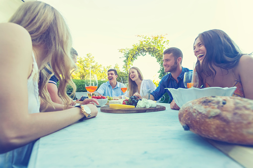 Group of young people eating outdoors. They are all happy, having fun, smiling laughing and talking. There are fruit and cheese platters on the table and some salad. Low angle view