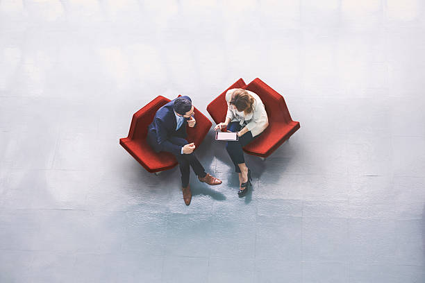 Overhead view of two business persons in the lobby A high-angle view of a businessman and a businesswoman sitting in the office building lobby and using a tablet computer generic description photos stock pictures, royalty-free photos & images