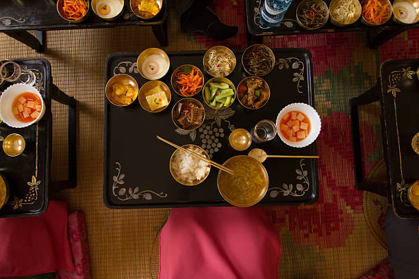 A vibrant overhead view of a wooden dining table laden with a variety of small bowls and plates of Korean food. The spread includes a hot stone bowl with rice, kimchi, sauces, and a large platter of fresh leafy greens. In the center, a person's hands are in the process of assembling a food wrap, capturing the communal and hands-on dining experience.