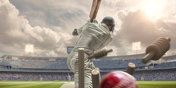A close up rear view image of a high speed cricket ball in mid air hitting stumps behind a cricket batsman during an outdoor game of cricket. The action takes place in a summer evening under a cloudy sky in a floodlight stadium full of spectators. The location is fictional.