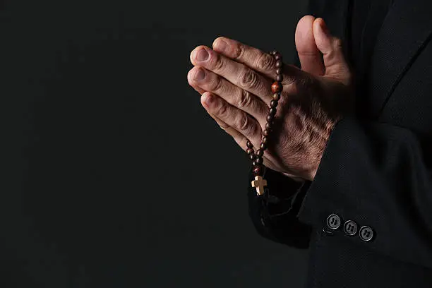 Hands of priest holding rosary and praying over black background