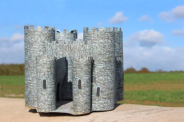 Photo showing a homemade cardboard castle, complete with battlements, turrets and crenellations.  This miniature medieval castle was made for a school project out of card and paper, and lots of glue, and is pictured on a wall, against a blue summer's sky.   A green field, countryside hedgerow and trees also form the distant background