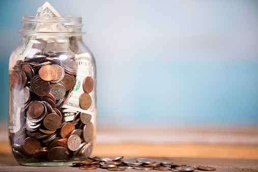 Money Jar filled with US currency: coins and paper money.  Savings or donations concept. Blue window background. 