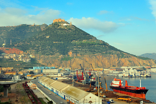 Oran, Algeria / Algérie: harbor scene - Bassin d'Arzew - warehouses and freighters unloading steel bars, harbour authority building and railway line - Djebel Murdjadjo mountain and the Santa Cruz fortress in the background - photo by M.Torres 
