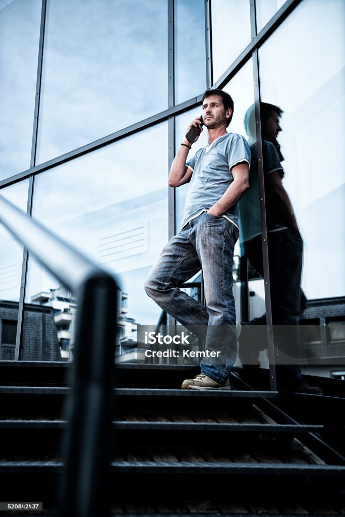 man talking on cellphone at modern office building Handsome man standing outdoors on the stairs of a modern office building leaning at the glass facade. He is talking at his cell phone with a hand in his pocket looking at view. Men Stock Photo