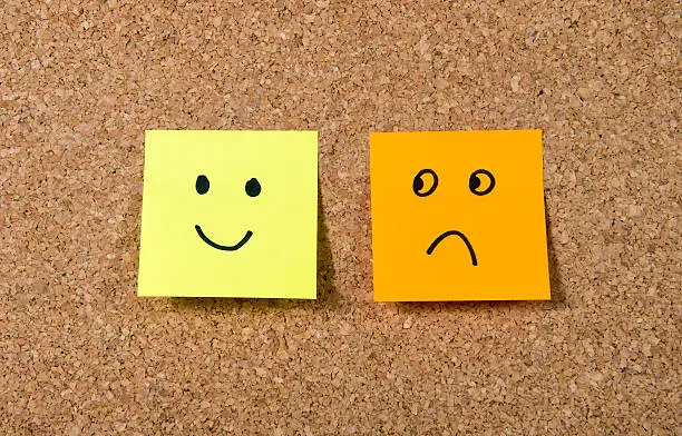 Two notes stuck on cork board or message board with smile and sad cartoon face expression in happiness versus depression and smile against adversity concept.