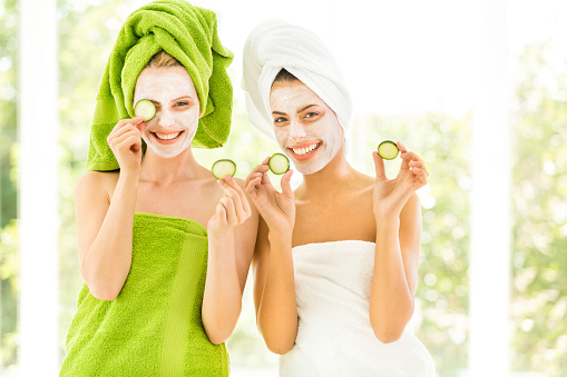 Two smiling young women with towel on head and slice of cucumber in hands.