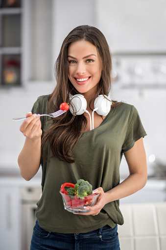 Portrait of a brunette woman while standing in the kitchen and holding a bowl with vegetables. She is wearing white headphones around her neck and holding a fork with cherry tomato.