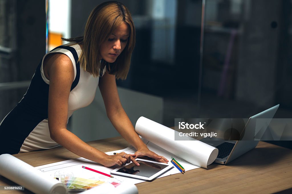 Architect using digital tablet late in her office. Architect designer reviewing blueprint in the office and using digital tablet. Businesswoman stay late in the office to finish project. Working late. Manager Stock Photo