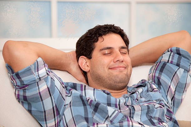 One handsome, Latin descent man happily napping or taking a mental break at home on living room sofa. Smiling, daydreaming, serene. Window background. Hands behind head. 