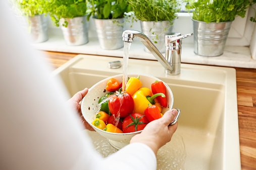 Housewife preparing vegetables for cooking in a domestic kitchen. She is washing paprika, tomatoes in a colander under running tap water. The water is splashing into the sink.