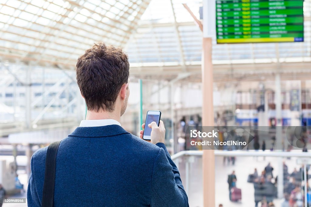 Traveler sending text message on smartphone in train station Traveler sending text message on smartphone with train timetable in background Train - Vehicle Stock Photo