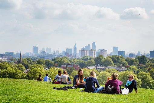 London, UK - September 15, 2014: Groups of friends sitting on a grassy hill at Primrose hill in London, UK, sharing food, stories and looking at the view. London Skyline and a partly cloudy sky in the background.