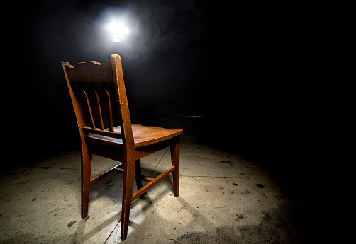 Isolated wooden chair in a dark scary prison with an interrogation spotlight.  The room looks like a dungeon or a prison cell where prisoners are interrogated.  The chair is empty and surrounded by dark shadows to give a feeling of terror and anxiety.  Some smoke is visible from the spotlight.