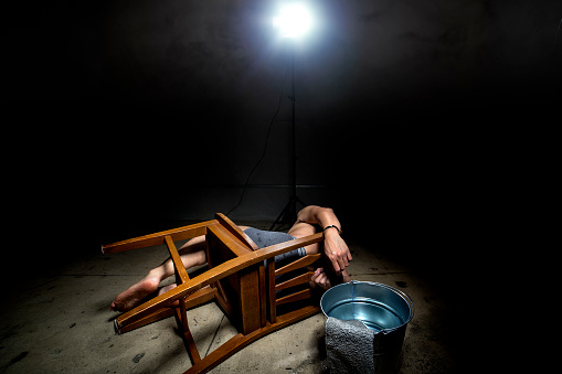 Prisoner being punished with cruel interrogation technique of waterboarding.  Beside him is a bucket of water used in the controversial torture technique.  The image depicts an illustrative editorial of the cruel political policy.  The man's identity is obscured.