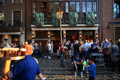 Dusseldorf, Germany - October 18, 2014: View on the popular brewery Uerige in Düsseldorf Altstadt with people drinking the so called Altbier (German for 'old beer'), a beer specialty of the city of Dusseldorf.  A waiter, called Köbes in the Rhineland, is passing the scene with a tablet of Altbier in the bottom left.