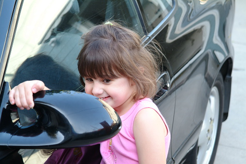 A two year old adorable little girl admiring herself in the drive side mirror of a sports car.
