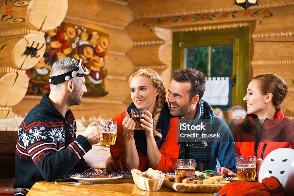 Friends having lunch and drinking beer after skiing Two adult couples wearing winter clothes sitting in a mountain restaurant after skiing, talking, enjoying dinner and drinking beer.  Adult Stock Photo